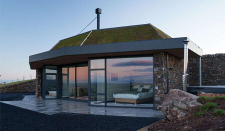 A modern stone and turf-roofed house catches the evening light, its glazed walls reflecting the sky above outdoor seating inside.