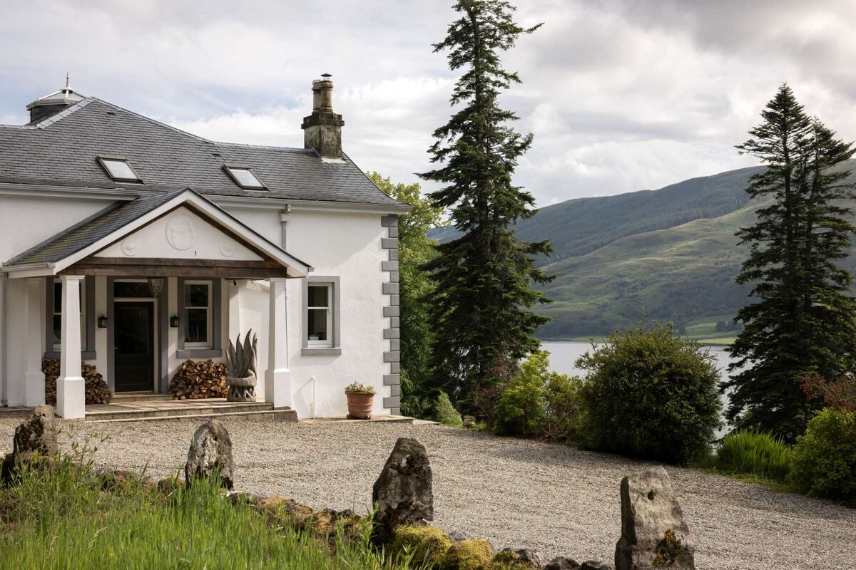 The porch and front door of Glenstriven House beside a gravel drive, with tall firs and Loch Striven behind.