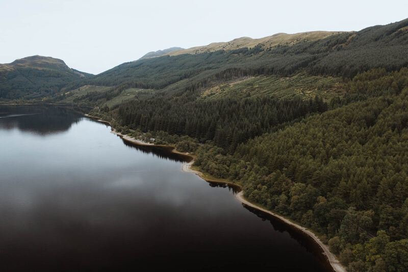 Forested hillside curves along a still loch beneath distant mountains