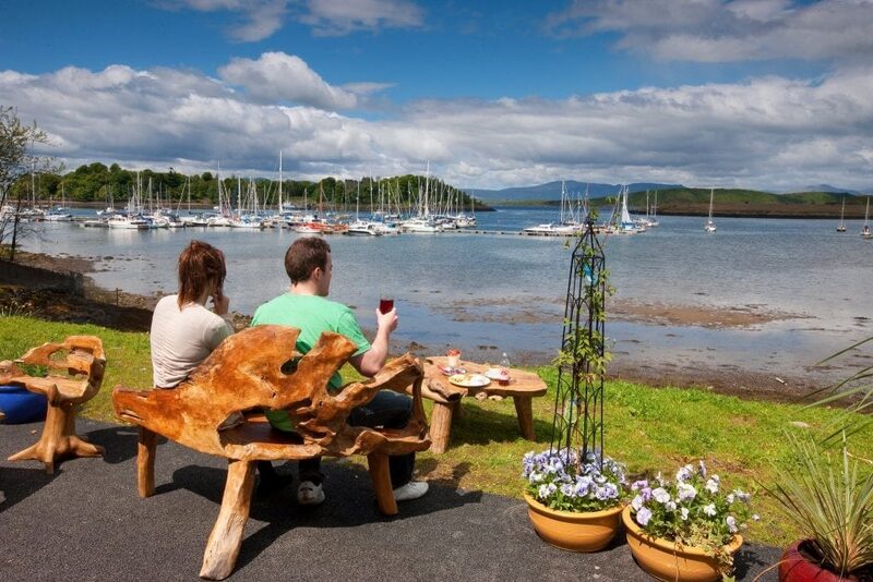 Couple share wine at a driftwood table overlooking a yacht-filled bay