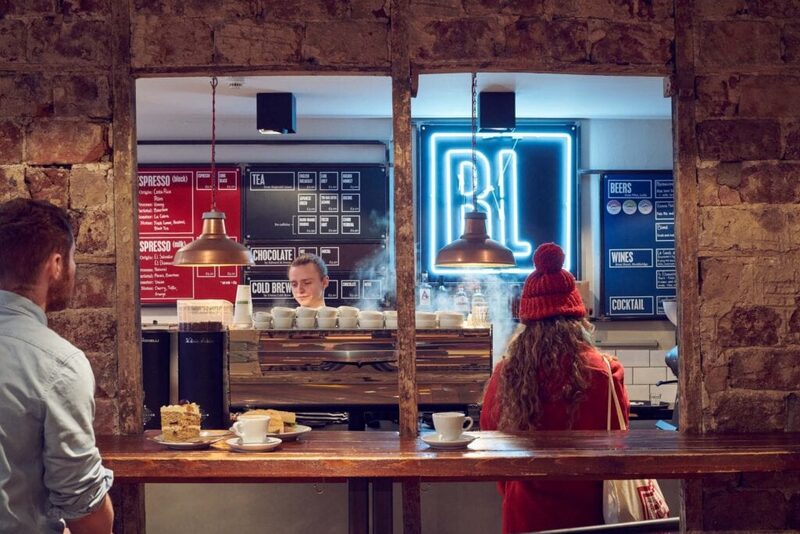 Barista serves coffee beneath a neon "BL" sign at a stone-walled café counter