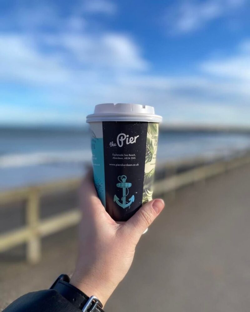 Hand holding a "The Pier" coffee cup above Aberdeen beach promenade