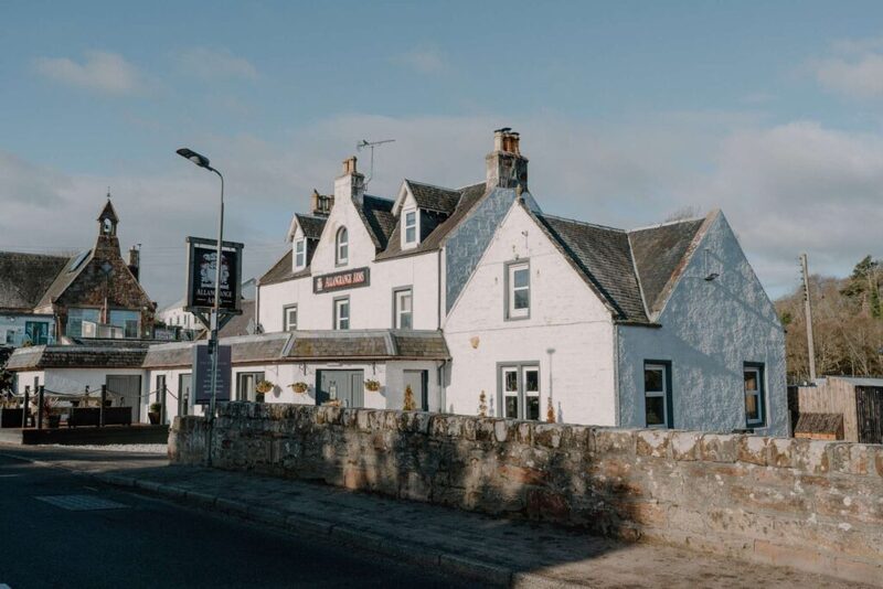 White-harled Clachan Inn with crow-stepped gables behind a stone roadside wall