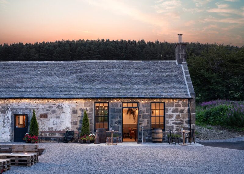 Stone cottage with lit doorways and slate roof at dusk, forest behind