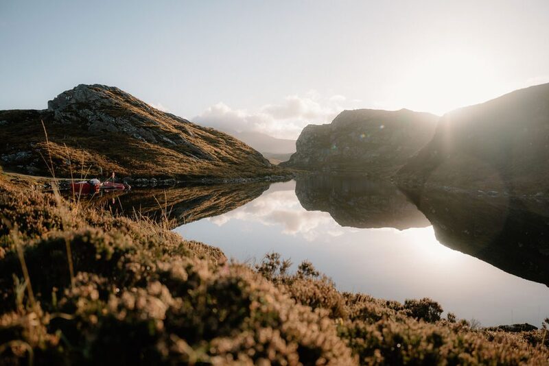Sunlight breaks between hills over a still loch with a red tent pitched at the shore
