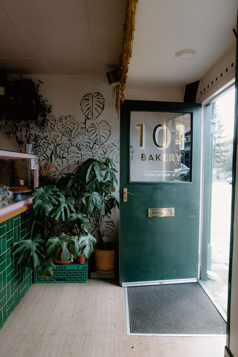 Green door reading "101 Bakery" beside leaf murals and potted plants