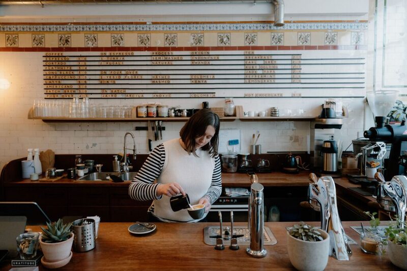 Barista in a striped jumper pours coffee beneath a chalked menu board