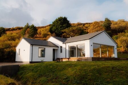 A white bungalow with a glazed gable end sits before an autumn hillside, a wooden bench on the lawn in front.