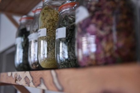 Glass jars of dried herbs and flowers lined up on a wooden shelf
