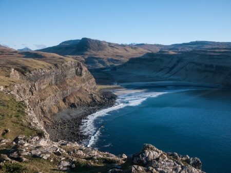 Cliffs and shingle beach curve around a blue sea inlet under distant hills