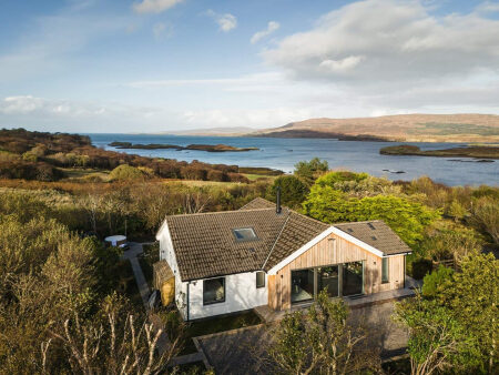 An aerial view shows a white and timber-clad house set among autumn trees above a bay dotted with small islands.