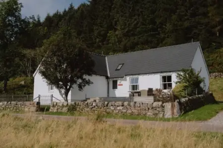 A white cottage with a grey slate roof stands behind a stone wall among trees on a hillside.