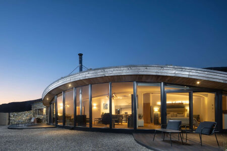 A curved timber-roofed house glows with lit windows at dusk, chairs set on the gravel and decking outside.