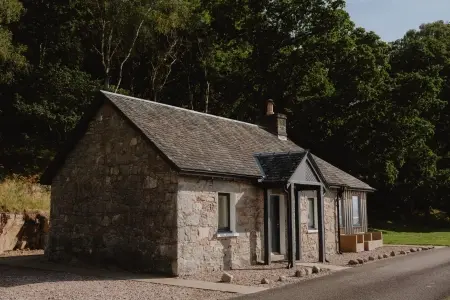 A single-storey stone cottage with a slated roof stands beside a road, mature trees rising behind.