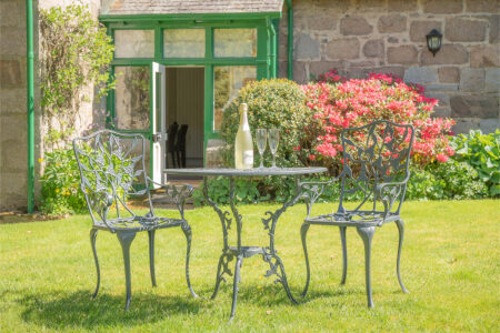 A round metal garden table with two chairs stands on a lawn beside a bottle of wine and glasses, pink flowering shrubs and a green-framed conservatory behind.