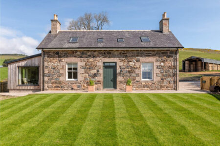 A stone farmhouse with a slated roof and green door sits behind a lawn, a timber garden room extending to one side.