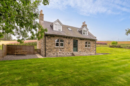 A stone farmhouse with dormer windows stands beside a picnic bench within a wooden fence enclosure.