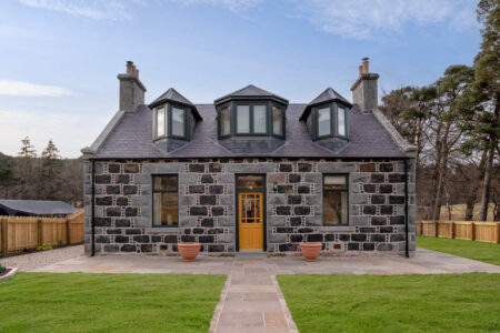 A stone house with three dormer windows and a yellow front door sits behind a lawn, terracotta pots flanking the entrance.