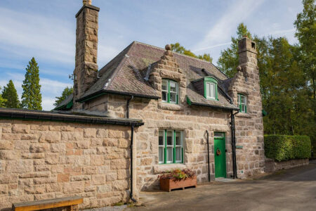 A stone gate lodge with steep gables and green-framed windows sits beside a stone boundary wall, a planter of flowers by the green door.